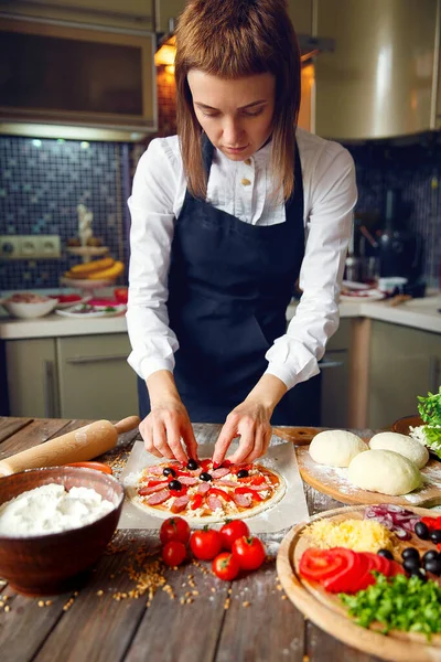 Woman in white shirt and apron putting the ingredients on the pizza. Pizza concept. Production and delivery of food