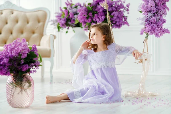 Menina bonita da idade pré-escolar em um vestido delicado sentado atrás de um balanço decorado com flores de lilás — Fotografia de Stock