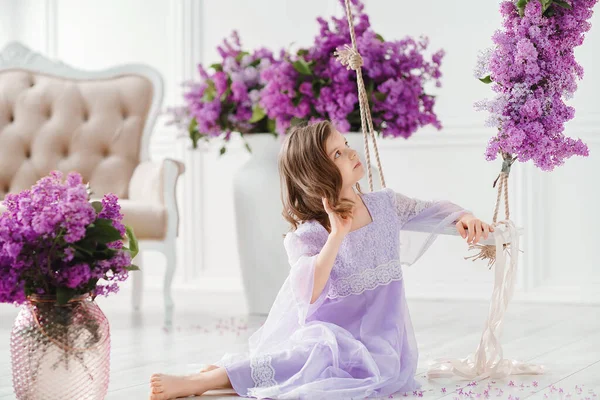 Hermosa niña de edad preescolar en un vestido delicado sentado detrás de un columpio decorado con flores de color lila — Foto de Stock