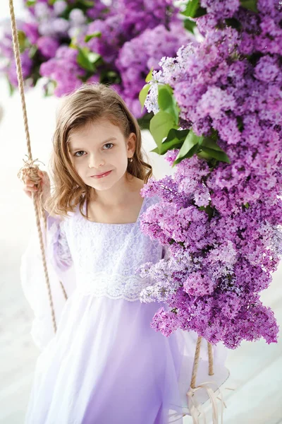 Hermosa niña de edad preescolar en un vestido delicado sentado en un columpio decorado con flores de color lila — Foto de Stock