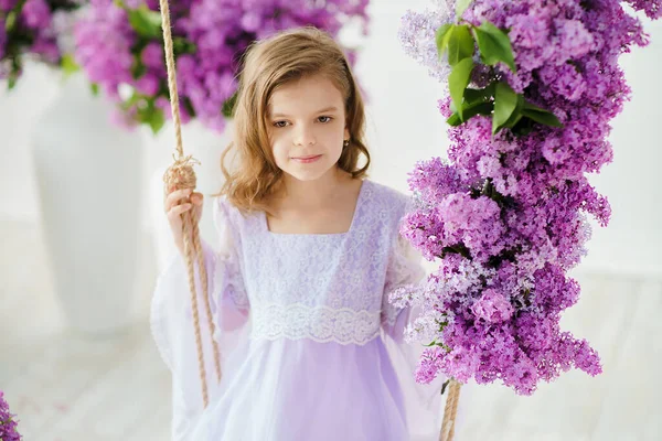 Vacker liten flicka i förskoleåldern i en delikat klänning sitter på en gunga dekorerad med blommor av syren — Stockfoto