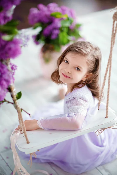 Красивая маленькая девочка дошкольного возраста в нежном платье сидит на качелях, украшенных цветами сирени — стоковое фото
