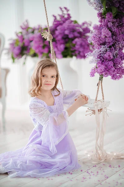 Schöne kleine Mädchen im Vorschulalter in einem zarten Kleid sitzt auf einer Schaukel mit Fliederblumen dekoriert — Stockfoto