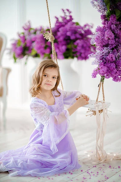 Schöne kleine Mädchen im Vorschulalter in einem zarten Kleid sitzt auf einer Schaukel mit Fliederblumen dekoriert — Stockfoto