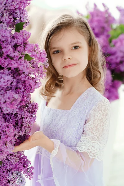 Красивая маленькая девочка дошкольного возраста в нежном платье сидит на качелях, украшенных цветами сирени — стоковое фото