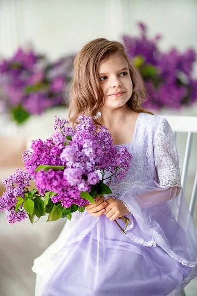 Beautiful little girl of preschool age in a delicate dress sitting on a chair behind swing decorated with flowers of lilac — Stock Photo, Image