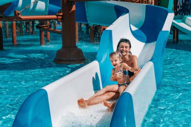 Mayo giymiş küçük bir kız mavi slaytlardan havuza iner, anne ve kız açık havuzda oynar ve yüzer.