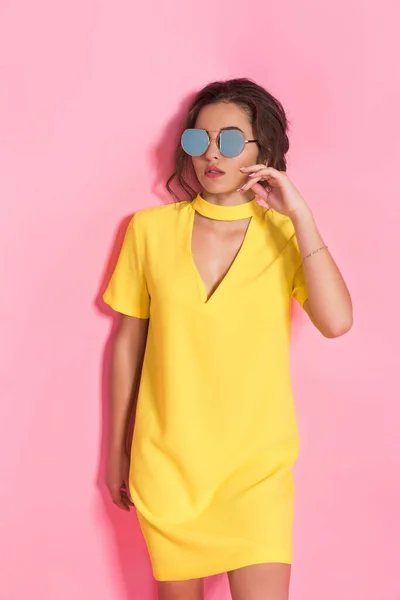 Красива дівчина в жовтій сукні та сонцезахисних окулярах позує на рожевому фоні в студії — стокове фото