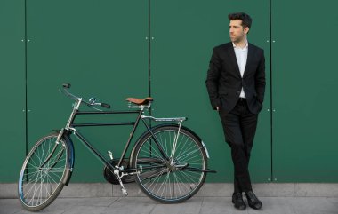 Takım elbiseli adam klasik bisikletiyle yeşil duvara yaslanıyor.