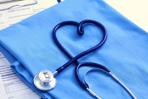 Medical stethoscope twisted in heart shape lying on patient medical history list and blue doctor uniform closeup. Medical help or insurance concept. Cardiology care, health, protection and prevention — Stock Photo, Image