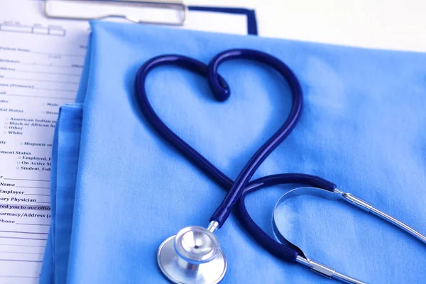 Estetoscopio médico retorcido en forma de corazón que se encuentra en la lista de antecedentes médicos del paciente y primer plano uniforme médico azul —  Fotos de Stock