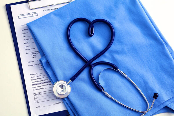 Медицинский стетоскоп скрученный в форме сердца, лежащий в списке пациентов и синей униформе врача крупным планом
