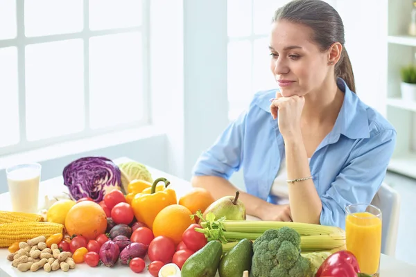 Feliz jovem dona de casa sentada na cozinha preparando alimentos de uma pilha de frutas e legumes orgânicos frescos diversos — Fotografia de Stock