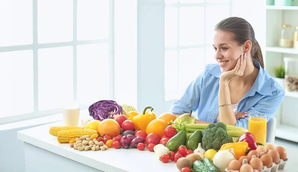 Happy young housewife sitting in the kitchen preparing food from a pile of diverse fresh organic fruits and vegetables — Stock Photo, Image