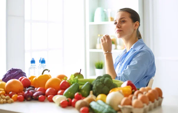 Feliz jovem dona de casa sentada na cozinha preparando alimentos de uma pilha de frutas e legumes orgânicos frescos diversos — Fotografia de Stock