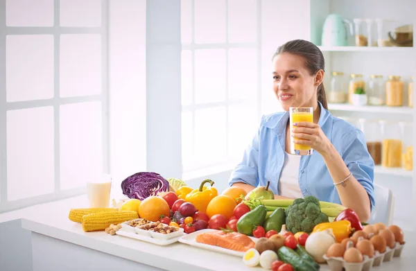 Happy young housewife sitting in the kitchen preparing food from a pile of diverse fresh organic fruits and vegetables — Stock Photo, Image