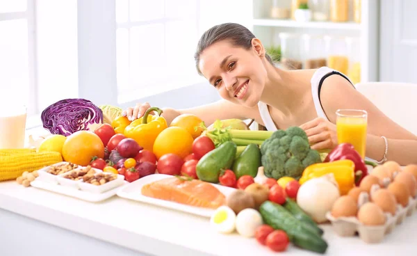 Feliz ama de casa joven sentada en la cocina preparando alimentos a partir de una pila de diversas frutas y verduras orgánicas frescas — Foto de Stock