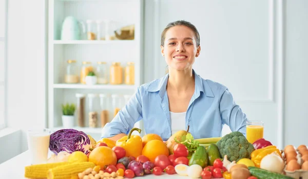 Feliz ama de casa joven sentada en la cocina preparando alimentos a partir de una pila de diversas frutas y verduras orgánicas frescas —  Fotos de Stock