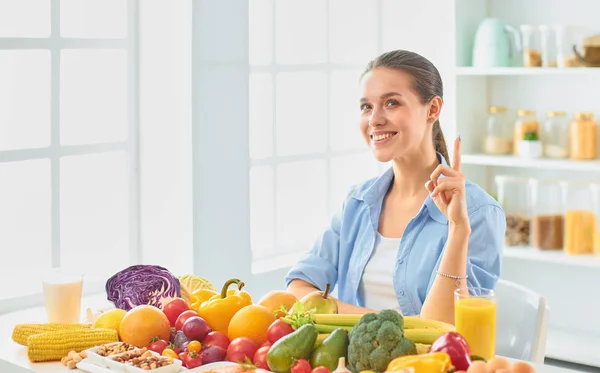 Happy young housewife sitting in the kitchen preparing food from a pile of diverse fresh organic fruits and vegetables — Stock Photo, Image