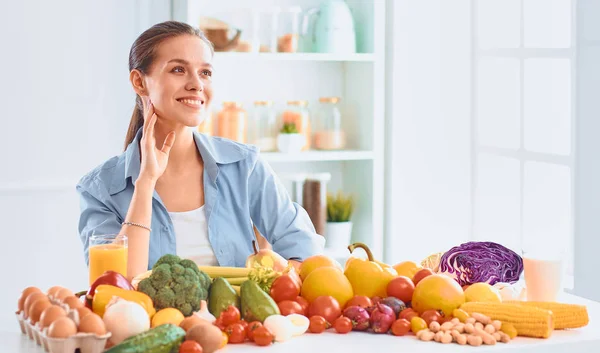 Feliz jovem dona de casa sentada na cozinha preparando alimentos de uma pilha de frutas e legumes orgânicos frescos diversos — Fotografia de Stock