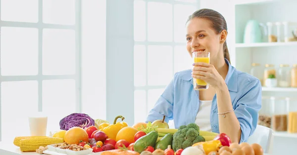 Happy young housewife sitting in the kitchen preparing food from a pile of diverse fresh organic fruits and vegetables, selective focus — Stock Photo, Image