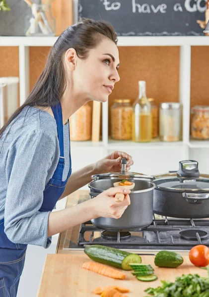 Hermosa chica está degustando comida y sonriendo mientras cocina en casa — Foto de Stock