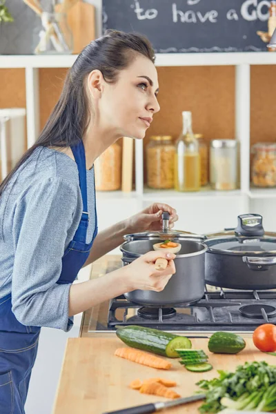 Hermosa chica está degustando comida y sonriendo mientras cocina en casa — Foto de Stock