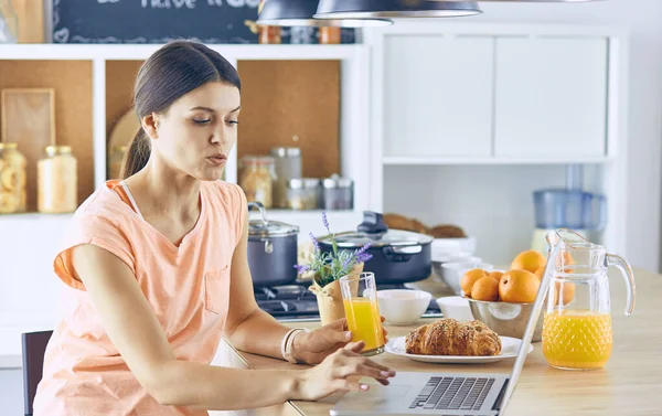 Uśmiechający się ładna kobieta, patrząc na telefon komórkowy i trzymając szklankę soku pomarańczowego, jedząc śniadanie w kuchni — Zdjęcie stockowe