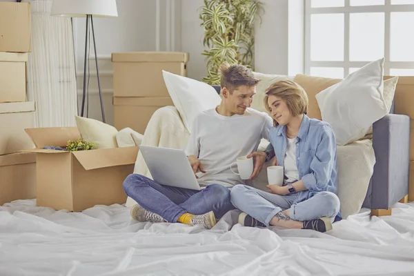 Retrato de pareja feliz mirando el ordenador portátil juntos sentados en una casa nueva, rodeados de cajas — Foto de Stock