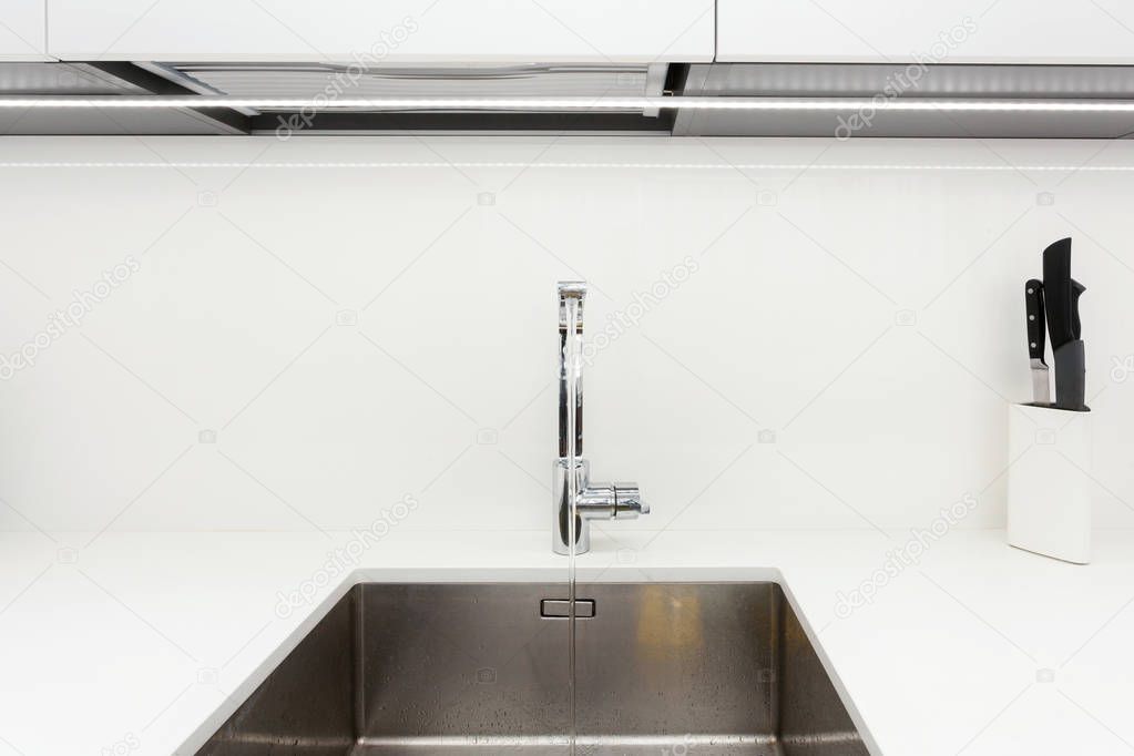 Modern designer chrome water tap over stainless steel kitchen sink. Interior of bright white kitchen.