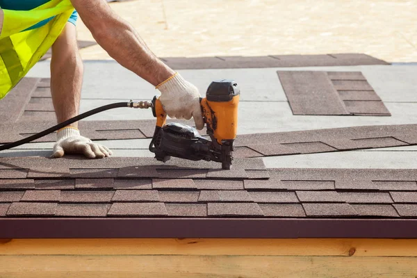 Construction worker putting the asphalt roofing (shingles) with nail gun on a new frame house. — Stock Photo, Image