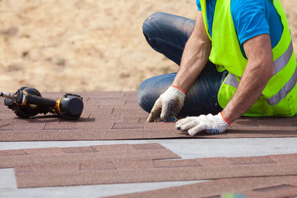 Roofer builder worker with nailgun installing Asphalt Shingles or Bitumen Tiles on a new house under construction.