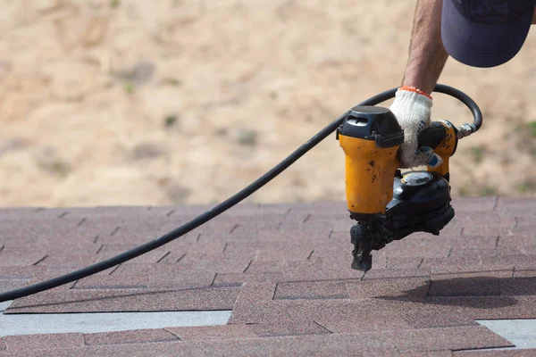 Roofer builder worker with nailgun installing Asphalt Shingles or Bitumen Tiles on a new house under construction. 