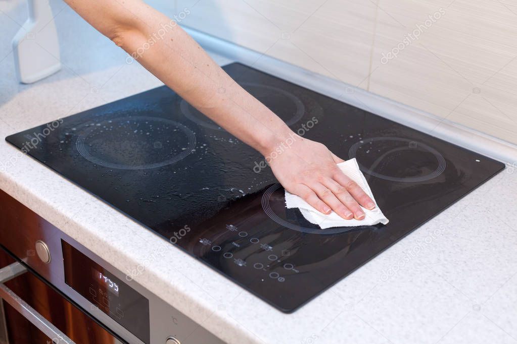 Housewife cleaning and polish electric cooker. Black shiny surface of kitchen top, hands, detergent.