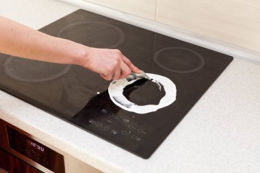Woman hands with bottle of cleaning detergent for induction stove applies a scour to the burner. clipart