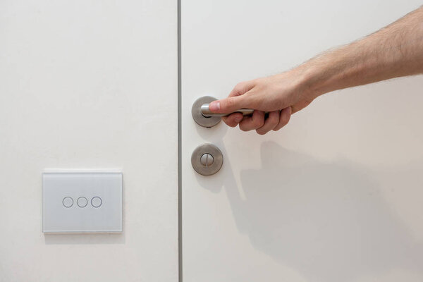Light switch next to the door handle. Open white door with metallic handle. The man opens the door