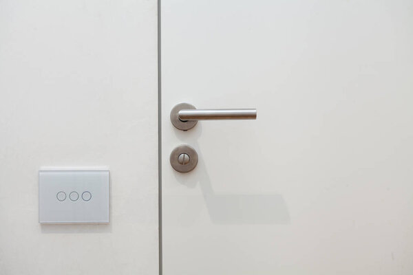 Light switch next to the door handle. Open white door with metallic handle.