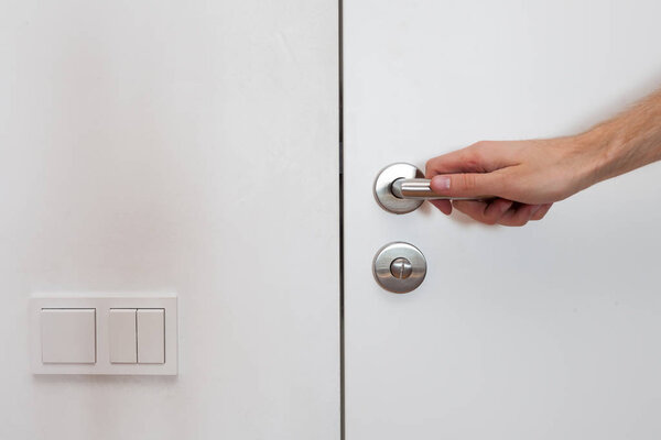 Light switch next to the door handle. Open white door with metallic handle. The man opens the door