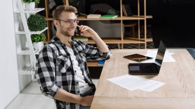 Masada modern bir dizüstü bilgisayarla oturan ve akıllı telefondan konuşan neşeli ve başarılı genç bir serbest yetişkinin panoramik görüntüsü