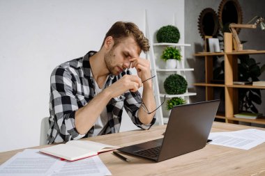 Uzun ve yorucu bir günün ardından çalışma alanında modern dizüstü bilgisayarın arkasında oturan yorgun ve bitkin bir genç iş adamı.