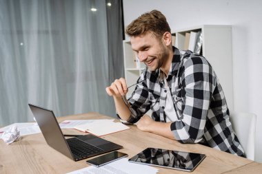 Ofiste oturan mutlu genç adam, modern dizüstü bilgisayar kullanıyor, müşteriyle online video görüşmesi yapıyor, geniş bir gülümseme sergiliyor.
