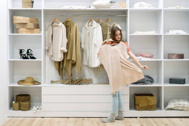 Çağdaş iç çamaşırlarıyla modern dolabın yanında duran genç bir kadın moda kıyafetlerini seçiyor, ne giyeceğini bilmemek, modaya uygun kıyafetler denemek gibi zor seçimler yapıyor.