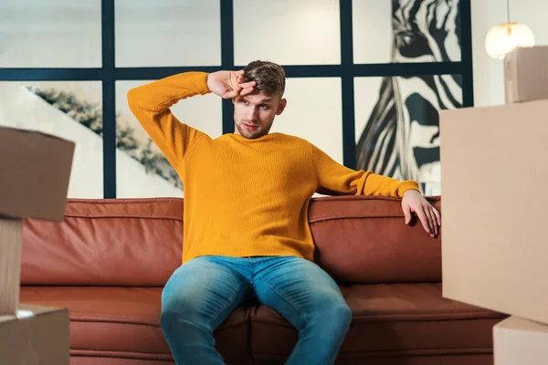 Tired and exhausted young adult man move in new rent house, sitting on couch, looking at boxes with stuff, resting and preparing unpacking belongings