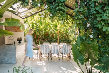 Yemek masasının yanında yürüyen bulanık bir kadın ve arka planda yeşil bitkilere karşı açık mutfakta sandalyeler.