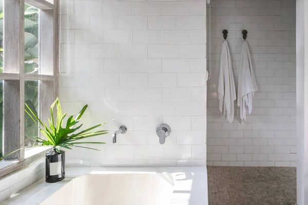 White bathroom interior, built bathtub, vase on top near wooden window frame with green plants on background, two dry, clean and fresh towel on wall hanger