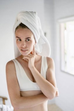 Dikey olarak gülümseyen yetişkin bir kadının aynaya bakıp tenini kontrol etmesi ve beyaz banyoda kafasında havluyla durması.