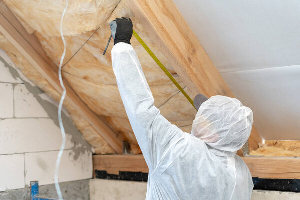 Back view of one unrecognizable workman in overalls working with rockwool insulation material, using measuring tape, standing inside new house under construction