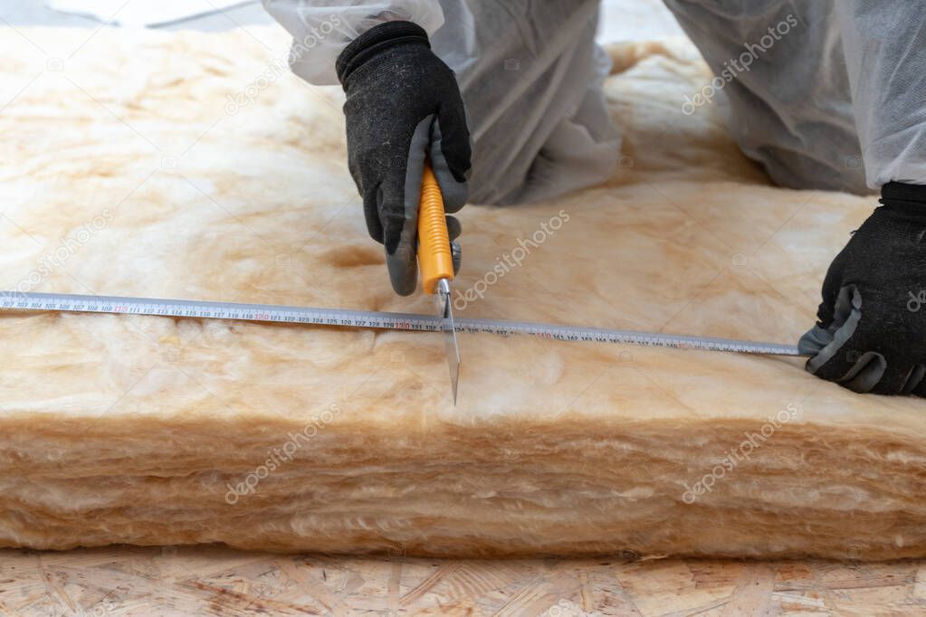 Cropped view of one professional worker in overalls working with rockwool insulation, cutting material, using knife tool and measuring tape, sitting inside new house under construction