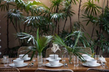 Otantik mutfakta, beyaz tabaklar, çatal bıçaklar ve vazoda yeşil dekorla süslü bir masa. Arka planda doğal bitkiler olan yemek odası.