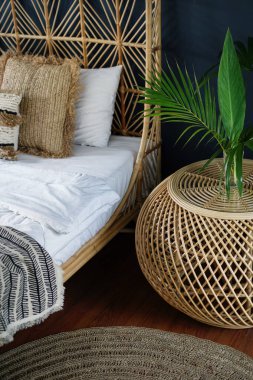 Etnik dekorlu, hasır komodinli, rahat yatak, halı, halı ve doğal yeşil bitki kompozisyonu olan dikey fotoğraf. Konforlu evde klasik yatak odası elementi